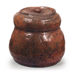 Ōhi I: gourd-shaped water jar, amber glaze Height: 19.0 cm, Mouth Diameter: 12.7 cm, Foot Diameter: 17.8 cm This is a gourd-shaped water jar with a tapered body, featuring a rough, hand-molded appearance. However, the craftsmanship around the rim and the incised lines running along the shoulders are quite exceptional. It appears to be modeled after a Shigaraki water jar; the amber glaze covering the entire surface is thin, revealing the sandy clay body beneath. It seems the amber glaze was applied specifically to highlight the interesting texture of this raw clay surface. The base has been flattened with a spiral-shaped scraper, leaving a narrow rim of folded clay around the edge. The body features large, wide vertical scrapes, and the inside of the base is coated not with amber glaze but with red Raku glaze. It is a style well-suited for wabi-cha.